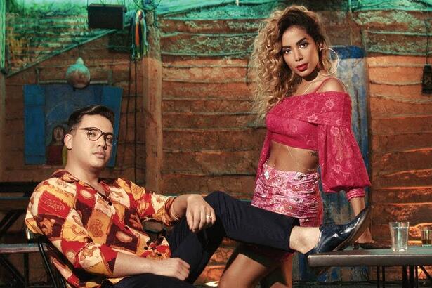 Wesley Safadão de camisa social estampada nas cores laranja, vermelho e branco, sentado, de óculos de grau, calça preta, com o pé esticado; Anitta com cabelo encaracolado, batom vermelho, saia rosa e tomara que caia em tom magenta, encostada numa mesa; ambos juntos para gravação de videoclipe