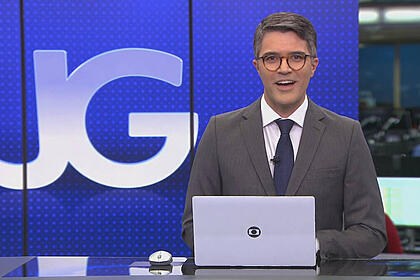 Bruno Tavares, de terno e gravata, sentado na bancada do Jornal da Globo, com o telão ao fundo