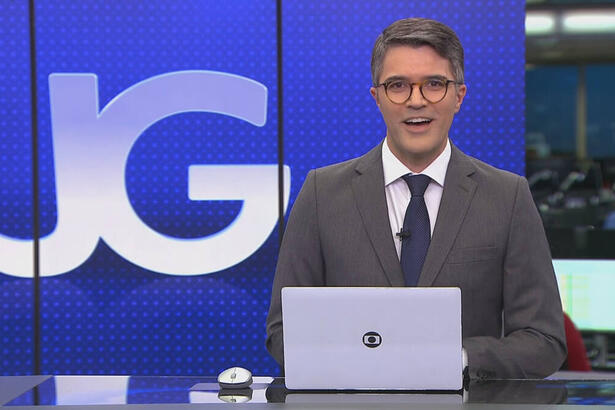 Bruno Tavares, de terno e gravata, sentado na bancada do Jornal da Globo, com o telão ao fundo
