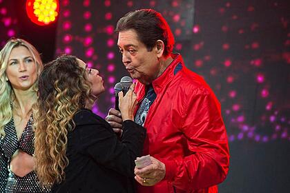 Daniela Mercury de blazer e top preto, puxando Faustão, de jaqueta vermelha, para dar um beijo, durante gravação de programa, na Band