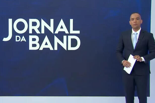 Diego Sarza, de terno, em pé em frente ao telão com o logo do Jornal da Band