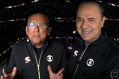 Galvão Bueno e Luís Roberto na cabine de transmissão da TV Globo no estádio Lusail, palco da final da Copa no Catar. Eles usam o uniforme de esporte da Globo e o estádio está com as luzes apagadas