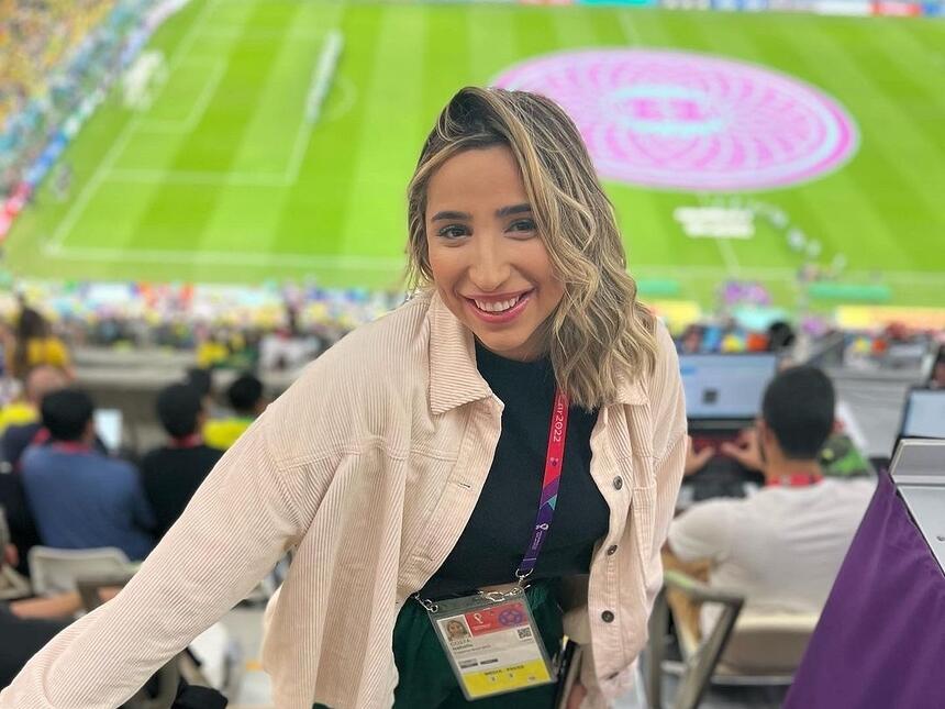Isabelle Costa de jaqueta bege camisa preta e crachá, de cabelo solto, sorrindo, no estádio