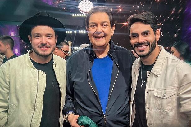 Israel de jaqueta bege e camisa preta sorrindo, de chapéu preto, ao lado de Faustão, de jaqueta preta e camisa azul, sorrindo, ao lado de Rodolffo, sorrindo, de jaqueta bege e camisa preta durante gravação do Faustão na Band