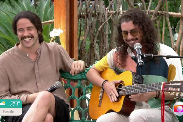 Juzé de óculos de sol, cabelo solto, segurando o violão; Lukete sorrindo, com cabelo preso, perna cruzada duraante gravação do É de Casa
