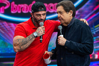 Kleber Bambam de camisa polo vermelha, chapéu preto, segurando o microfone com a mão direita, ao lado de Faustão, durante gravação do Faustão na Band