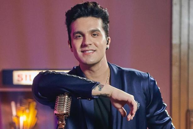 Luan Santana com camisa social manga longa azul, com o braço direito encostado no violão, sorrindo durante gravação de especial da TV Globo
