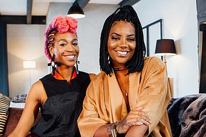 Ludmilla de roupão marrom, sorrindo, com cabelo trançado, ao lado de Karol Conk´´a, com cabelo rosa, sorrindo, de roupa preta, sentadas num sofá