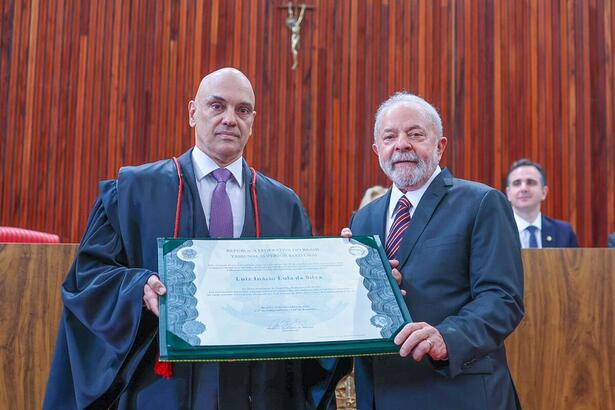 Lula e Alexandre de Moraes de blazer preto e camisa social branca, com gravata estampada vermelha, segurando diploma em cerimônia no TSE