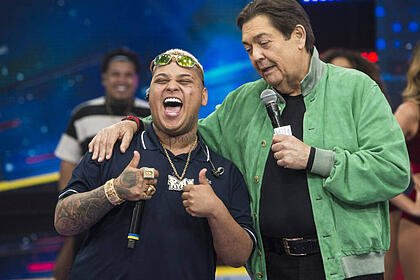 MC Ryan SP sorrindo, com a boca aberta, cabelo loiro, segurando o microfone com a mão direita; Faustão de jaqueta verde, camisa preta, segurando o microfone com a mão esquerda durante gravação de programa