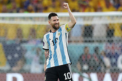 Messi com o sorrio aberto, em campo com a camisa da argentina, em jogo da copa do mundo. Ele está com o braço esquerdo levantado