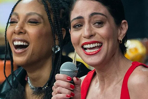 Carol Castro de regata vermelha, sorrindo, de batom vermelho, cabelo umido, ao lado de Negra Li, de look preto, sorrindo durante gravação do Faustão na Band
