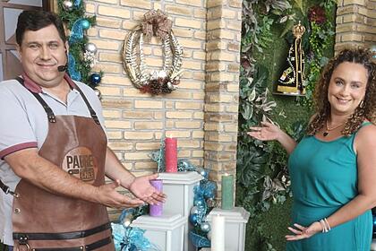 Padre Chef Evandro de avental marrom, de couro, apontando com as mãos para o lado esquerdo, sorrindo, de camisa cinza;  Letícia Andrade de macaquito verde, apontando com as mãos para a direita, em pé, ao lado do Padre Chef; ambos em pé, durante gravação do 'Sabor de Vida', da TV Aparecida