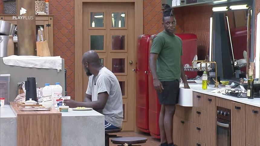 Pelé em pé na cozinha olhando para André que está sentado na mesa.