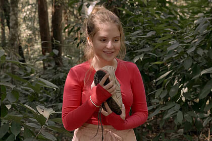 Atriz sophia valverde, como poliana, em trecho da novela poliana moça. Ela usa uma roupa com tom rosa, abraça um pé de tênis, em uma mata