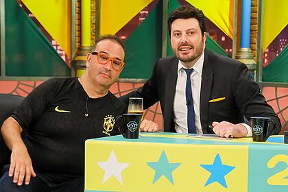 Renato Tortorelli de camisa da Seleção Brasileira preta, de óculos de grau, sentado num sofá branco ao lado de Danilo Gentili, durante gravação do The Noite, do SBT