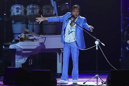 Roberto Carlos com blazer e calça azul claro, com as mãos abertas, cantando durante show no Rio de Janeiro