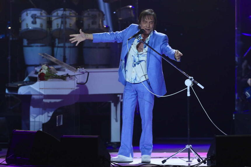 Roberto Carlos com blazer e calça azul claro, com as mãos abertas, cantando durante show no Rio de Janeiro