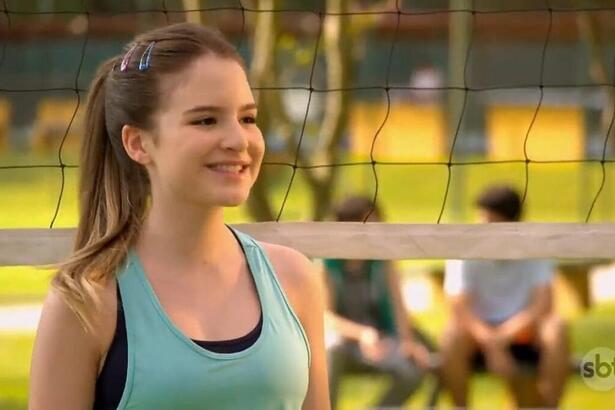 Sophia Valverde, como Poliana, em trecho da novela Poliana Moça. Ela está sorrindo, em frente a uma rede de vôlei em um campo