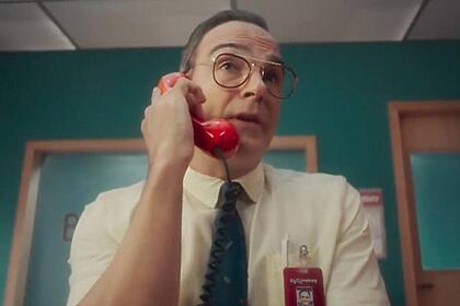 Tadeu Schmidt de calça bege, camisa social branca, gravata azul, segurando um telefone laranja num escritorio durante gravação de chamada para o BBB, da TV Globo