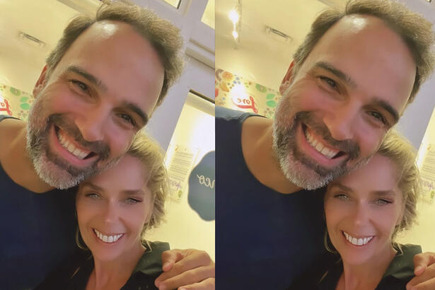 Adriane Galisteu sorrindo, de camisa social verde musgo, abraçada com Tadeu Schmidt, que está sorrindo, usando camisa básica preta;