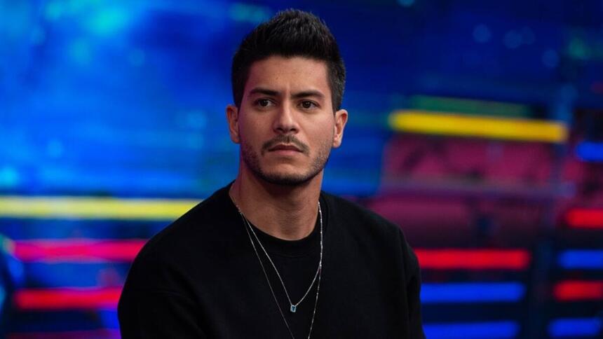 Arthur Aguiar de camisa manga longa preta, com um colar de prata, sentado, durante gravação de programa na TV Globo