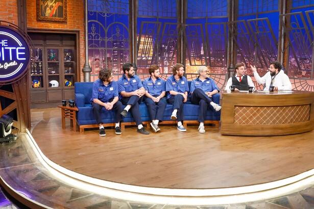 Banda Jovem Dionísio vestida de azul, sentados no sofá azul do The noite. Ao lado está os humoristas Léo Lins e Murilo Couto sentados na bancada apresentando o programa.