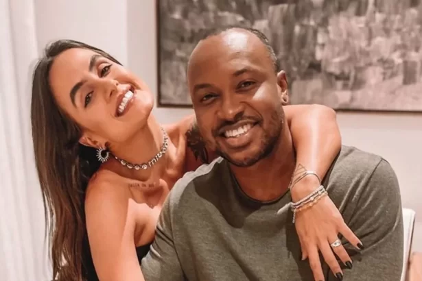 Carol Peixinho sorrindo, com a mão no pescoço de Thiaguinho, sentada num sofá