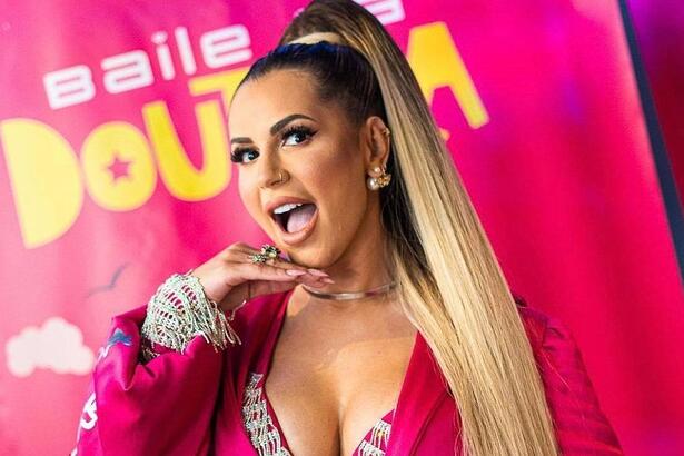 Deolane Bezerra com a mão no queixo, boca aberta, usando blazer decotado na cor rosa durante presença vip em festa