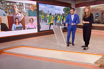 Erick bang e poliana abritta, em pé nos estúdios da globonews, durante a cobertura dos atos terroristas. Comentaristas aparecerem no telão
