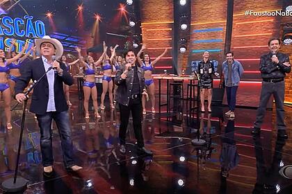 Cantores Chitãozinho e Xororó cantando no palco do Faustão Na Band. Ambos seguram seus microfones que estão apoiados em um pedestal. Ao fundo tem as bailarinas do programa com roupa roxa e ao lado tem os apresentadores da atração, Anne Lottermann, João Guilherme Silva e Fausto Silva.