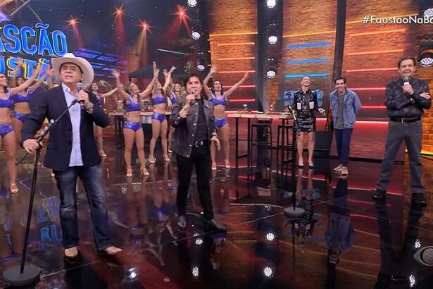Cantores Chitãozinho e Xororó cantando no palco do Faustão Na Band. Ambos seguram seus microfones que estão apoiados em um pedestal. Ao fundo tem as bailarinas do programa com roupa roxa e ao lado tem os apresentadores da atração, Anne Lottermann, João Guilherme Silva e Fausto Silva.