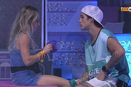 Bruno sentada segurando um copo de roupa brilhosa de frente para Gabriel que está sentado vestido com uma camisa azul e bermuda branca.