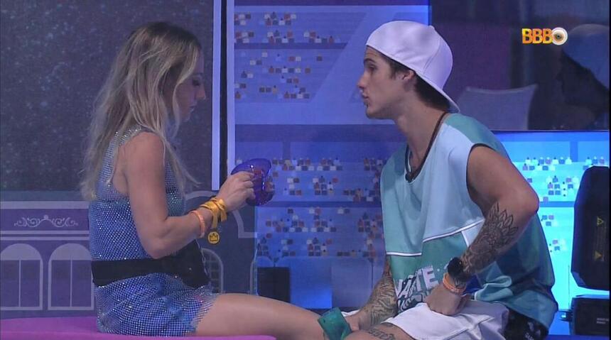 Bruno sentada segurando um copo de roupa brilhosa de frente para Gabriel que está sentado vestido com uma camisa azul e bermuda branca.