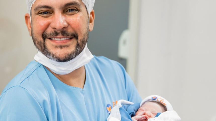 Jorge com a roupa de centro cirúrgico, com a filha recém-nascida no colo