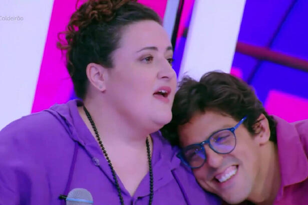 Ju Almeida de camisa roxa, segurando o microfone, sendo abraçada por Rafael Infante no Caldeirão, da TV Globo
