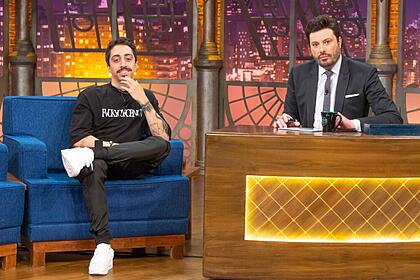 Lucas Inutilismo vestindo roupa preta com tênis branco, está sentando no sofá azul do The Noite apoiando uma mão no queixo e sorrindo. Ao lado está Danilo Gentili apresentador do programa com um semblante sério e terno e gravata com aos mãos apoiadas na bancada do seu talk show.