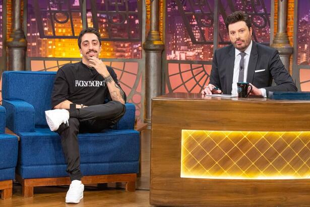 Lucas Inutilismo vestindo roupa preta com tênis branco, está sentando no sofá azul do The Noite apoiando uma mão no queixo e sorrindo. Ao lado está Danilo Gentili apresentador do programa com um semblante sério e terno e gravata com aos mãos apoiadas na bancada do seu talk show.