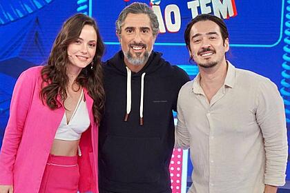 Marcos Mion vestido com roupa preta no meio dos seus convidados. Com ele está Marcos Veras vestido com roupa branca e Rosanne Mulholland com roupa rosa.