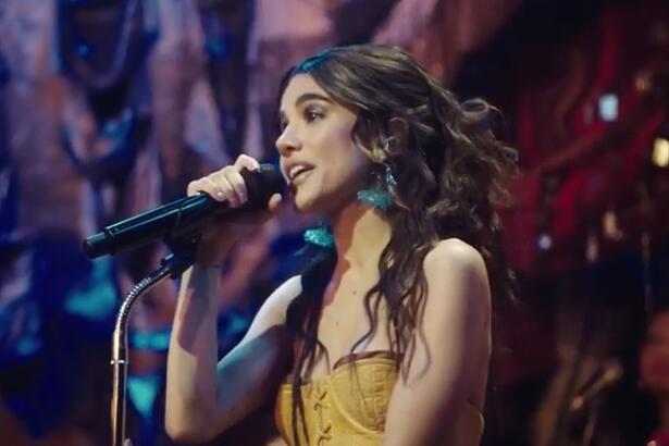 Manu Gavassi segurando um microfone, de vestido amarelo, cabelo ondulado, grande, solto, com brinco de fios azuis durante gravação do Acústico MTV