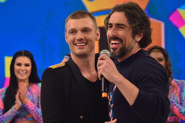 Marcos Mion segurando o microfone com a mão esquerda, sorrindo, com a mão no ombro direito de Nick, do Backstreet Boys, que está de blazer e camisa preta, sorrindo, durante gravação do Legendários, da Band