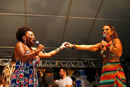 Margareth Menezes de vestido estampado em tons de ciano, segurando a mão de Ivete Sangalo durante show em Salvador