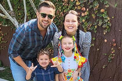 Mariana Bridi com vestido quadriculado e calça jeans, sorrindo, com tranças no cabelo; Rafael Cardoso com camisa quadriculada em tons de azul e branco, sorrindo, de oculos de sol, segurando os filhos durante passeio