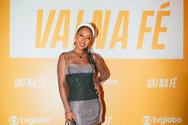 Cantora Negra Li com vestido de brilho cinza com preto sorrindo e apoiando a mão na cintura, com fundo de lançamento amarelo da novela 'Vai Na Fé' da TV Globo.