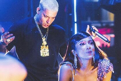 Filipe Ret de camisa preta e correntes no pescoço segura o microfone olhando para Anitta. Cantora está fazendo biquinho com a boca e mãos no joelhos durante um apresentação de um show.