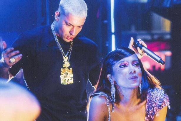Filipe Ret de camisa preta e correntes no pescoço segura o microfone olhando para Anitta. Cantora está fazendo biquinho com a boca e mãos no joelhos durante um apresentação de um show.