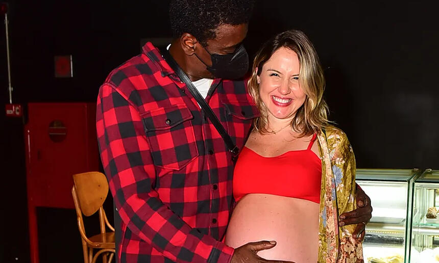 Seu Jorge de camisa quadriculada vermelha e preta, de mascara preta, com a mao esquerda na barriga de Karina durante evento em São Paulo