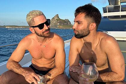 Thales Bretas de óculos de sol, short preto, segurando um drink ao lado de Silva, que está sem camisa, segurando uma taça de vinho, sentado num iate