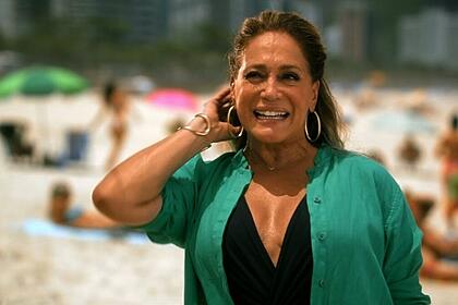 Susana Vieira de camisa preta e verde, com a mão na cabeça, sorrindo, durante gravação externa na praia