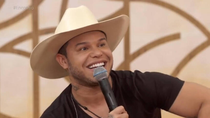 Tierry, com um chpéu branco na cabeça, sorrindo, com um microfone nas mãos, sentando no sofá do programa Encontro